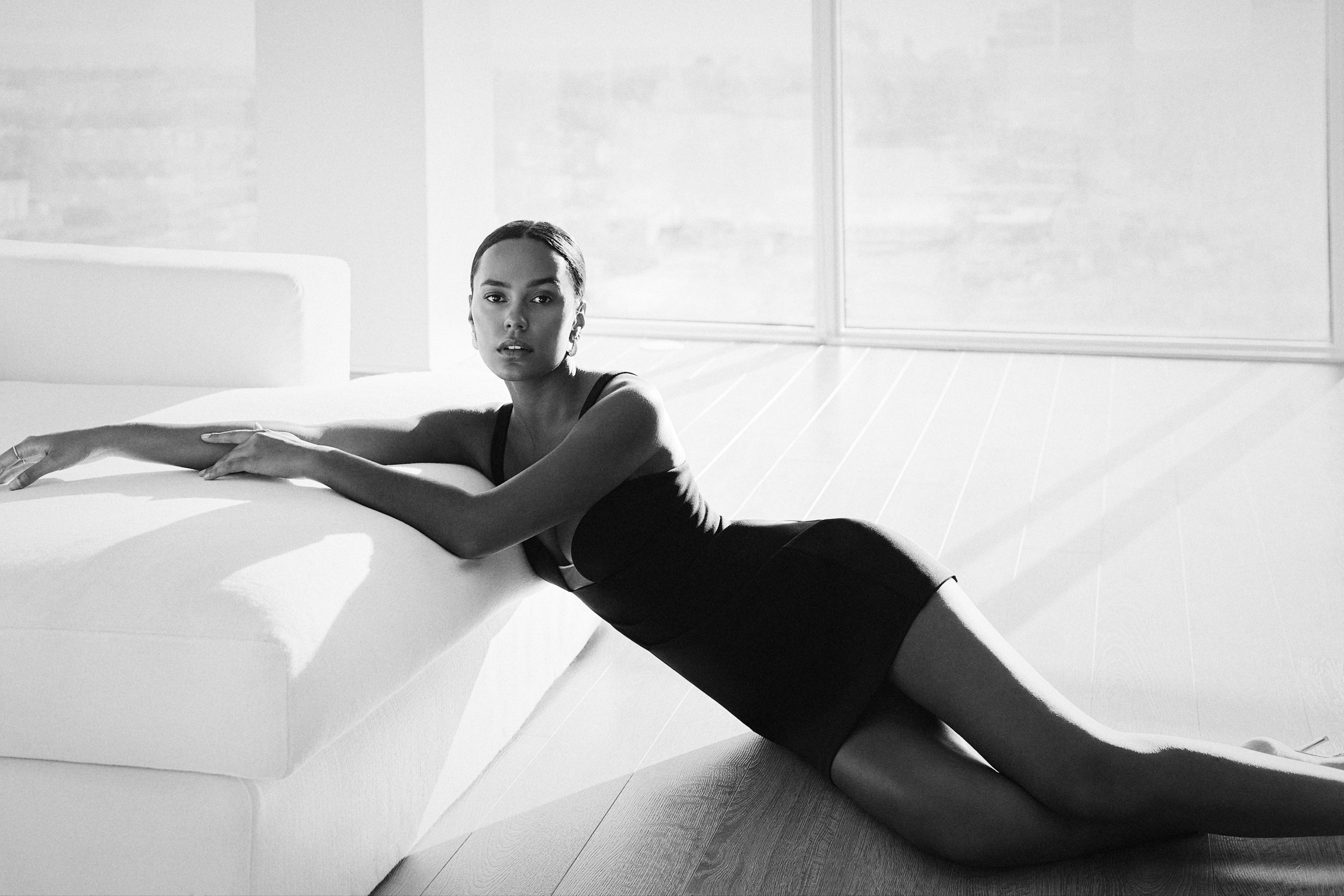 BCBGMAXAZRIA brand ambassador Ava Dash lying on the floor with her arms on a white couch in a black mini dress inside an apartment